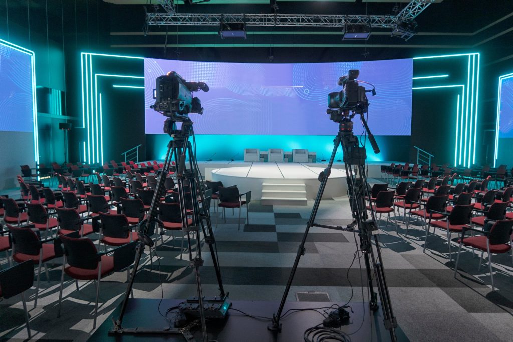 Two video cameras on tripods face a brightly lit stage with chairs—perfect for a corporate video production—in a modern auditorium accented by blue neon lights and a large digital screen backdrop.