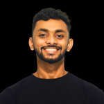 A young man with short curly hair and a beard smiles at the camera. He is wearing a plain black shirt, posed against a solid black background—an ideal look for modern corporate video production.
