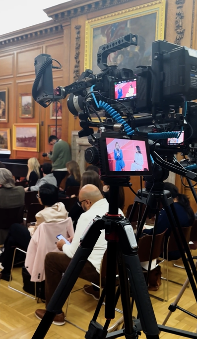 A video camera on a tripod records a speaker addressing an audience in a wood-paneled room adorned with framed paintings, capturing the essence of professional corporate video production. Some attendees watch the speaker while others glance at their phones.