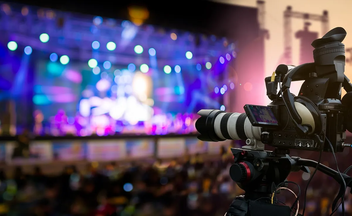 A professional video camera on a tripod is set up in the foreground, filming a brightly lit outdoor stage for a Corporate Video Production, with colorful lights and a blurred audience in the background.