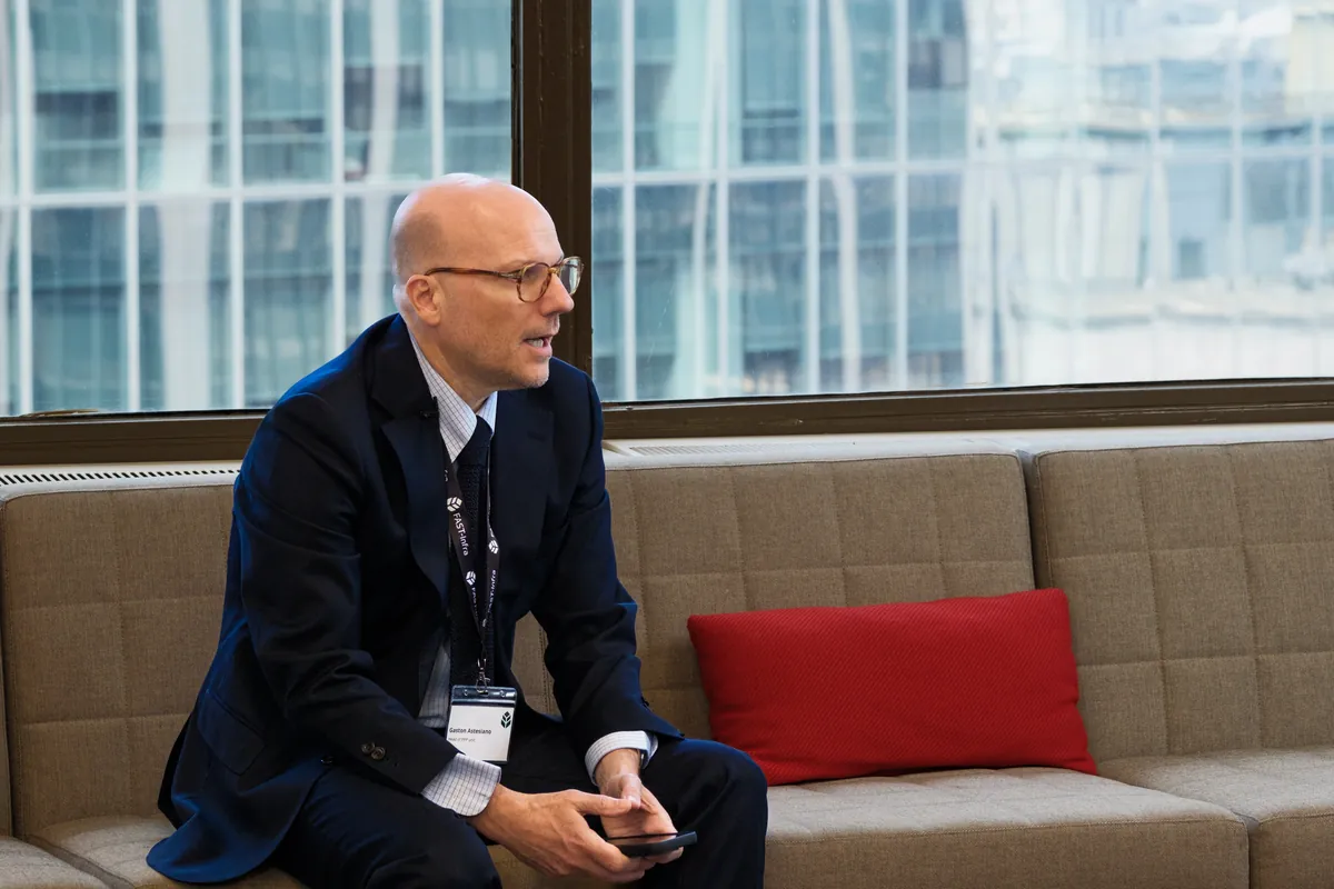 Man in a dark suit seated on a sofa during an interview setup near large windows