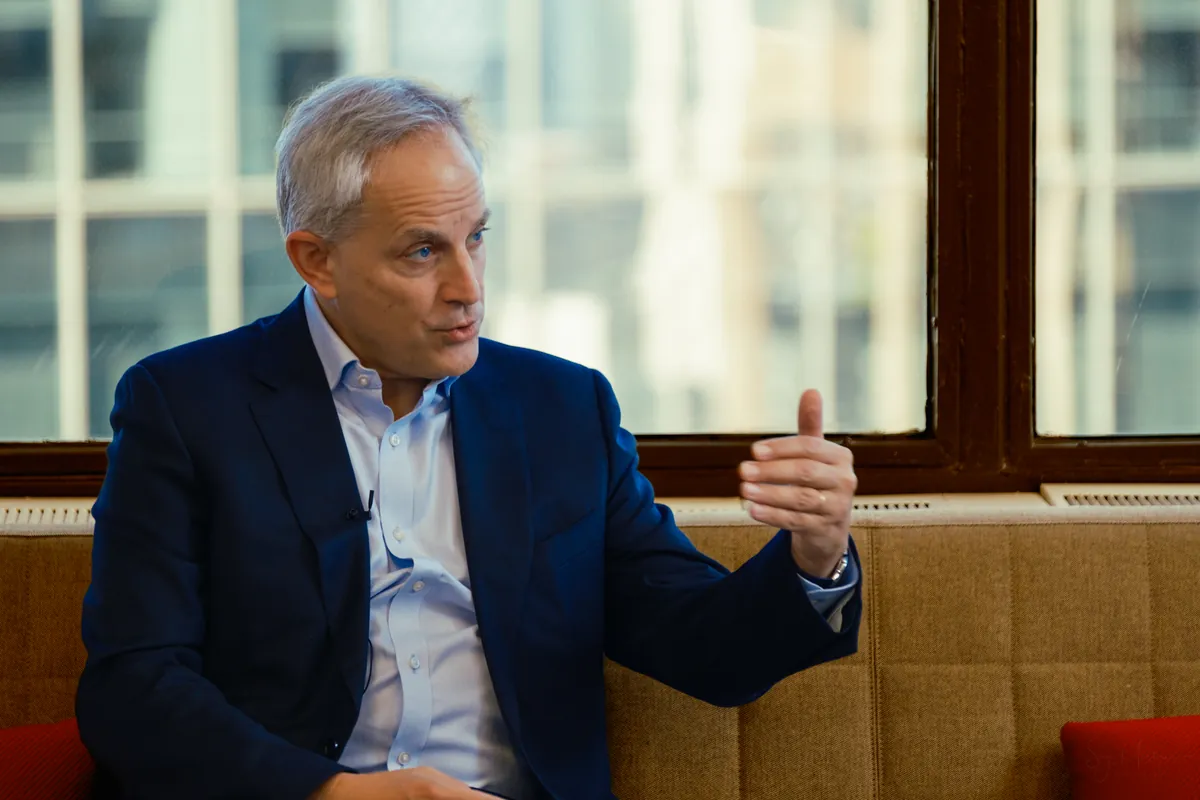 Man in a blue blazer gesturing while speaking during an interview on a sofa