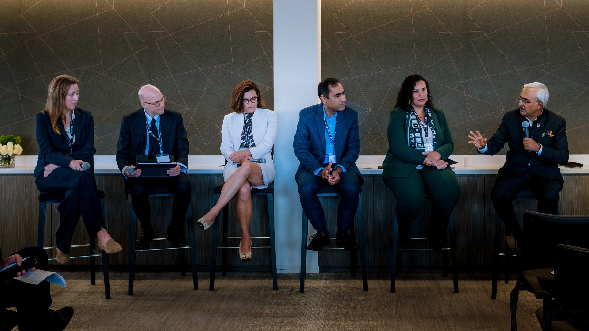 Six panelists seated on stools on a conference stage